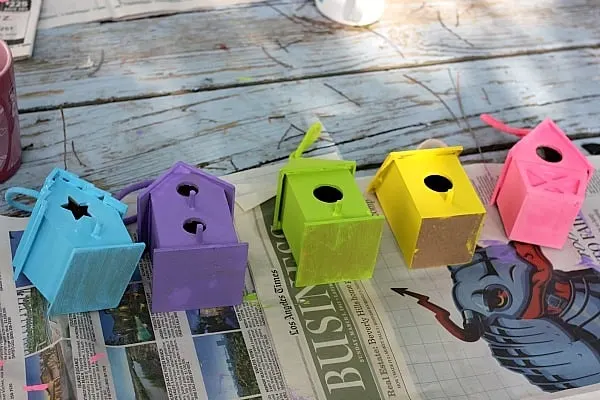 Five birdhouses painted with acrylic paint laying on newspaper drying 