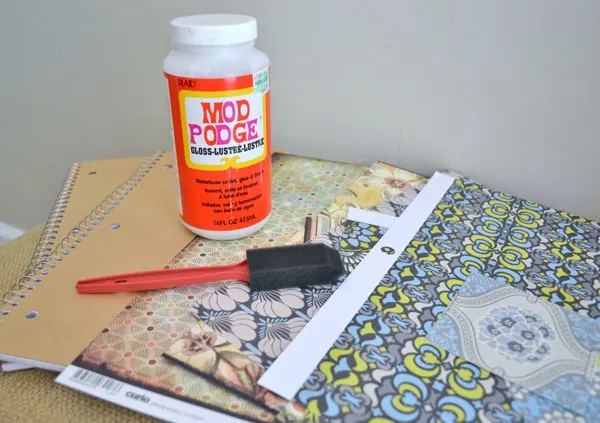 Bottle of Mod Podge, plain kraft notebooks, scrapbook paper, and a foam brush