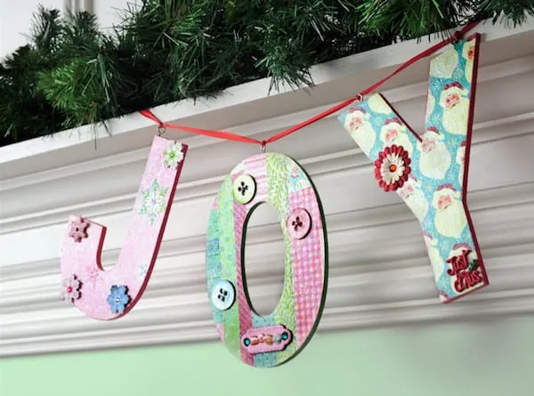 A decorative wooden banner spelling "JOY" hangs from a white fireplace mantle. The mantle is covered in fake pine needles.