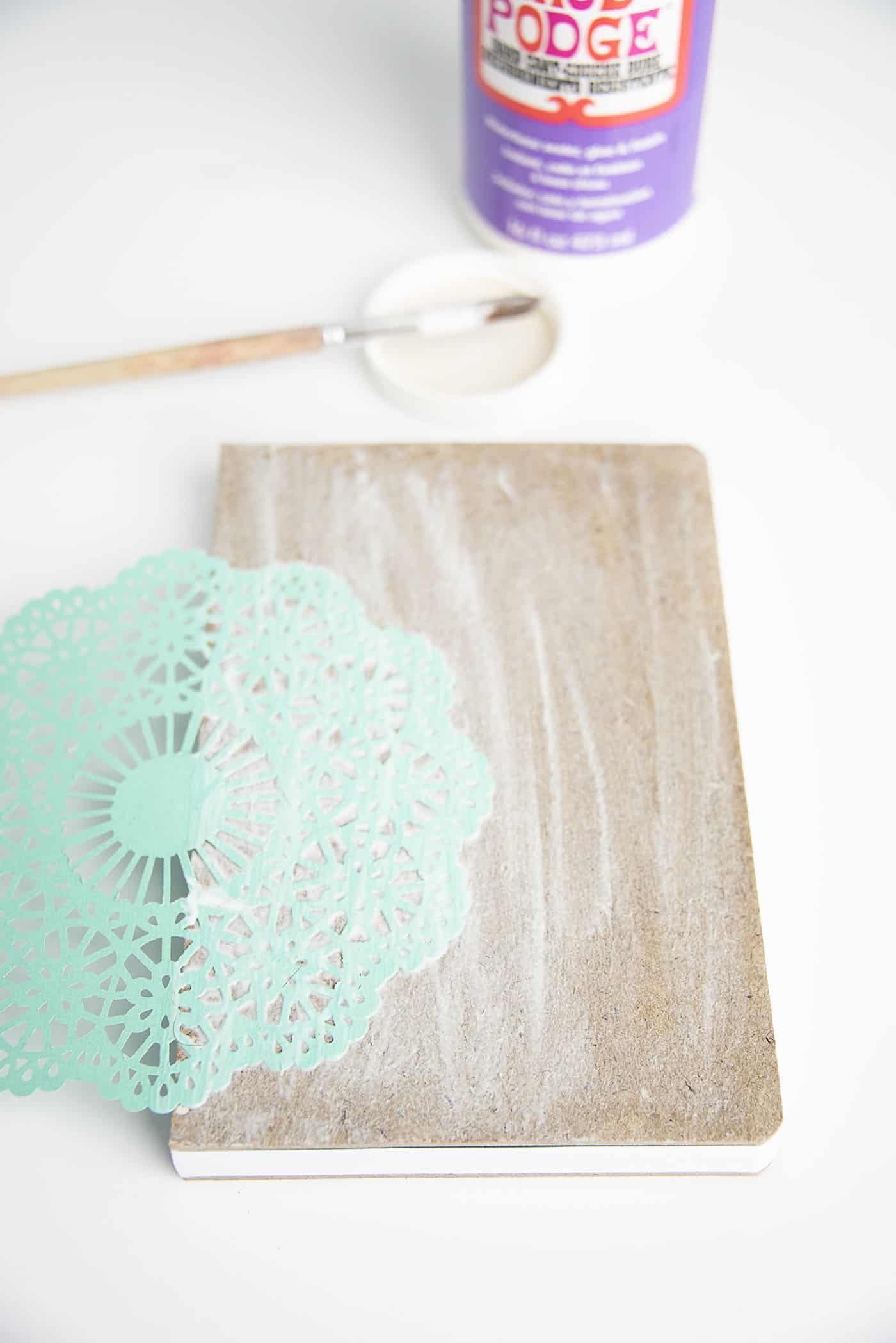 Adhering a paper doily to the front of a notebook with Mod Podge