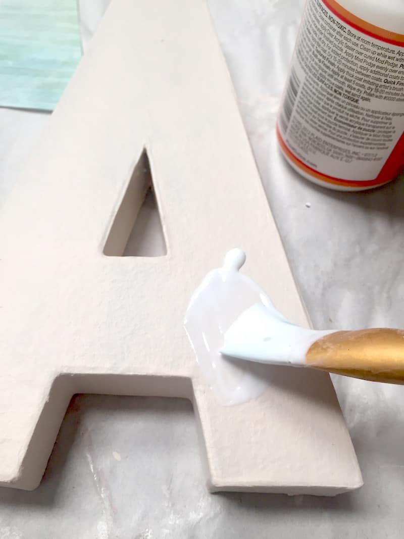 Applying Mod Podge to the top of a paper mache letter A with a paintbrush