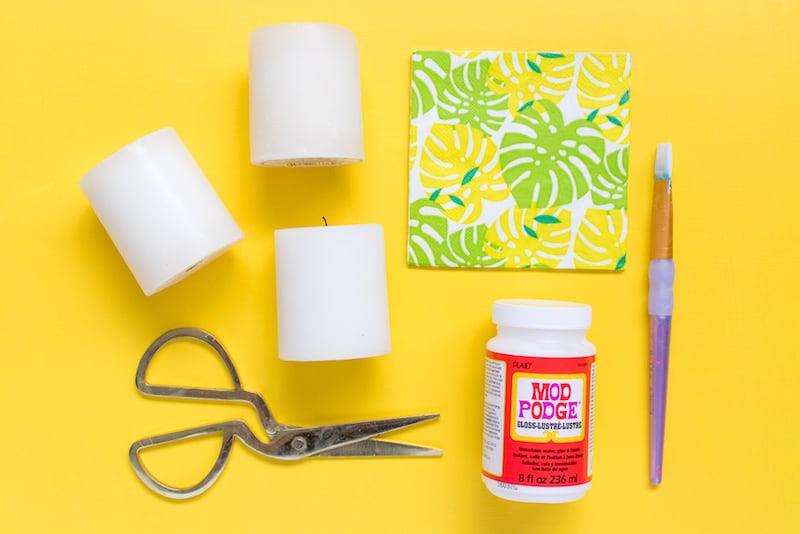 White candles, napkins, Mod Podge Gloss, paintbrush, and a pair of scissors on a yellow background