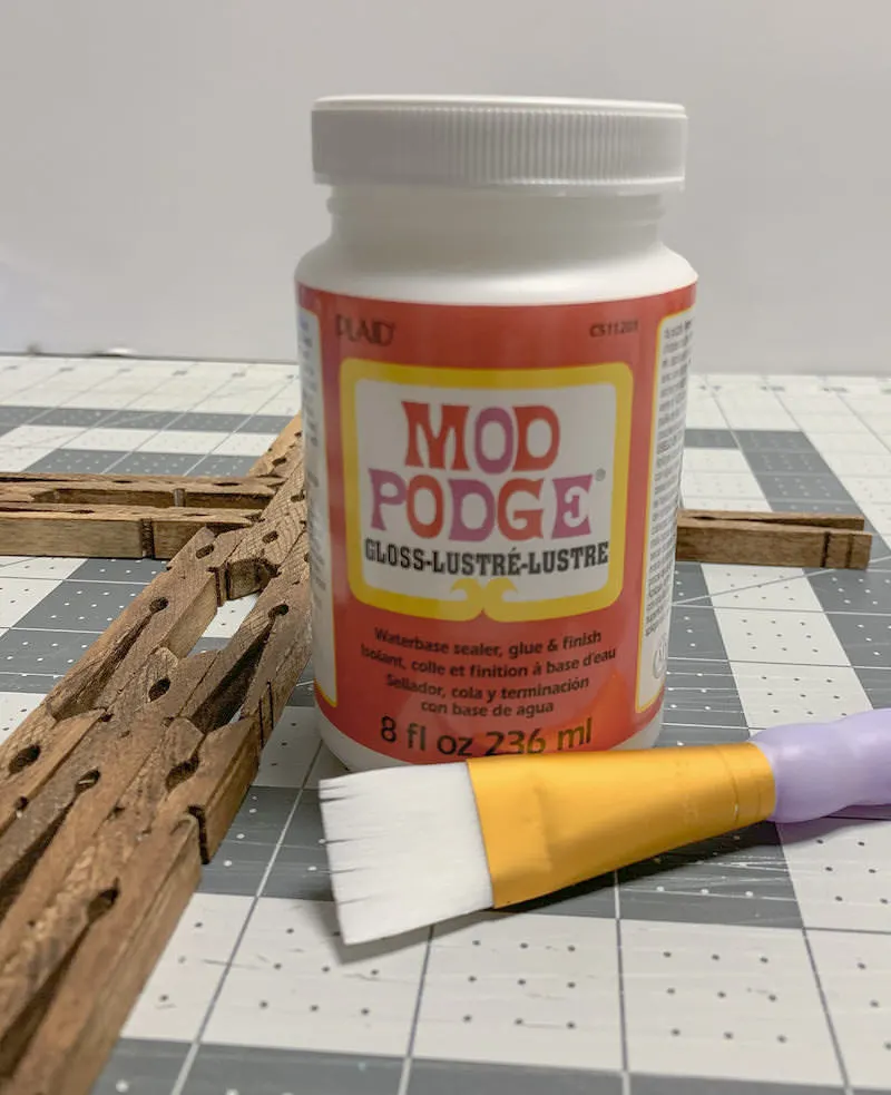 Bottle of Mod Podge next to a clothespin cross with a paintbrush