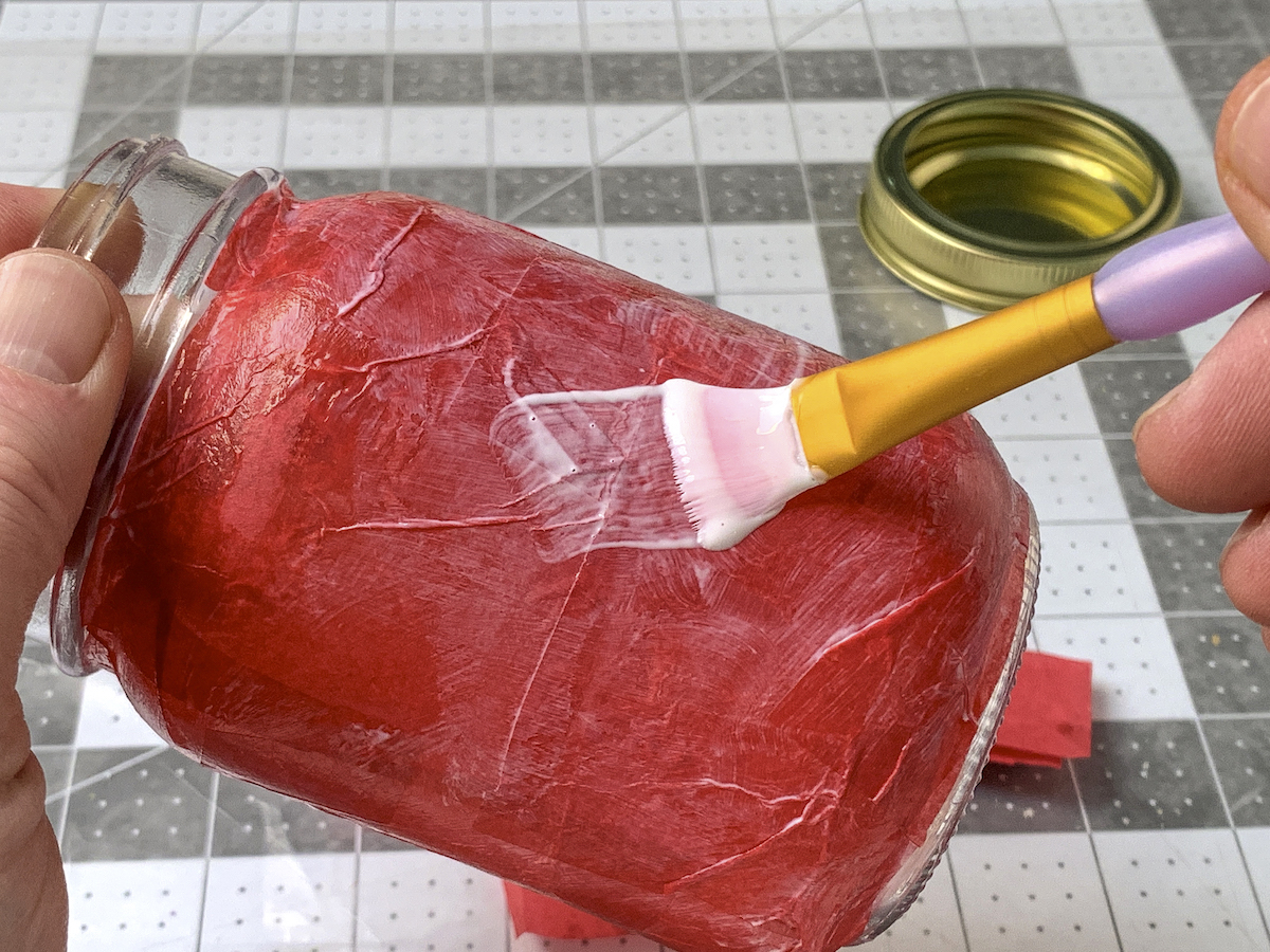Aplicação de uma camada final de Mod Podge em um frasco de vidro coberto com papel de seda