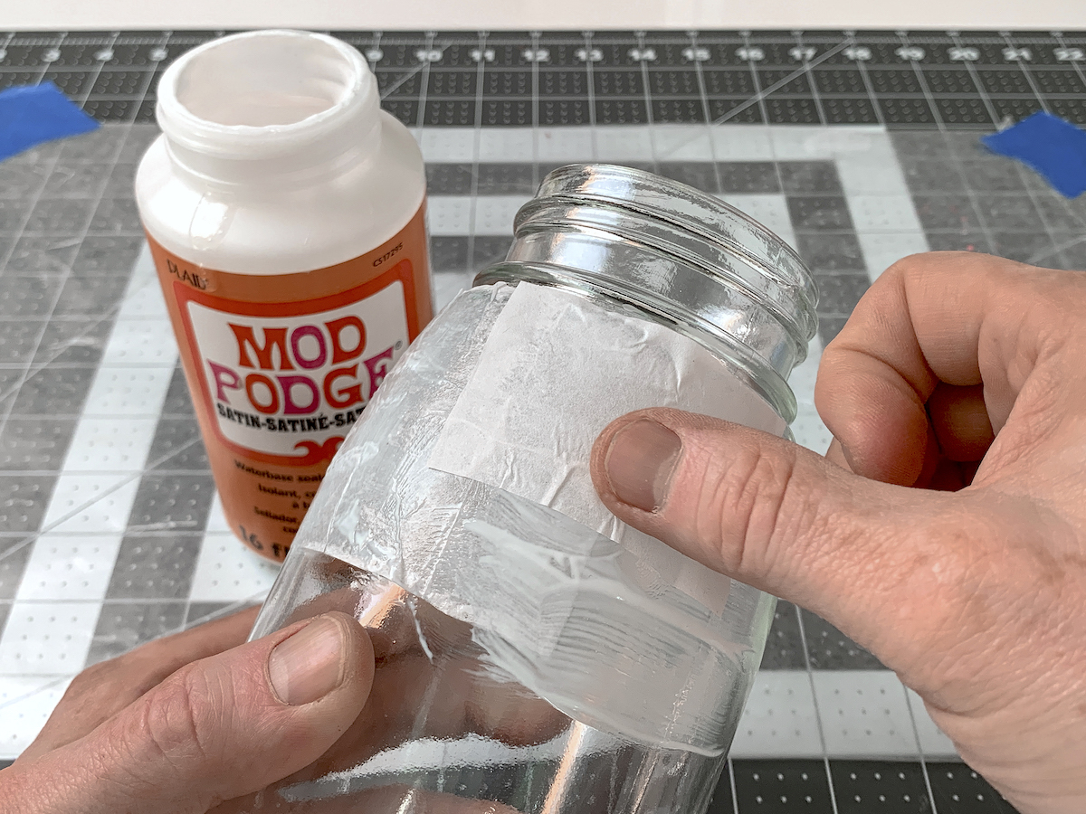 Hand placing white tissue paper down in wet Mod Podge on a jar